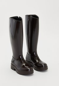 Botas negras brillantes de hasta la rodilla con suelas gruesas y cremallera lateral sobre un fondo blanco liso.