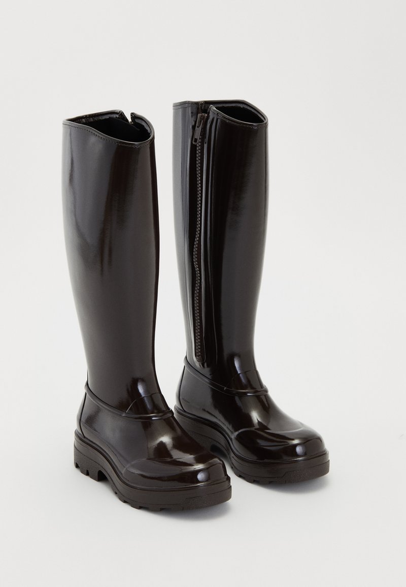 Botas negras brillantes de hasta la rodilla con suelas gruesas y cremallera lateral sobre un fondo blanco liso.