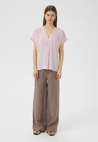 Blusa rosa chiaro a maniche corte con scollo a V e vestibilità ampia, abbinata a pantaloni larghi in lino marrone e infradito nere.