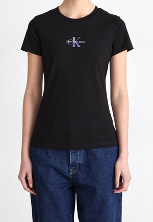 Henkilö pukeutunut mustaan istuvaan lyhythihaiseen t-paitaan, jossa on purppuranvärinen Calvin Klein Jeans -logo, yhdistettynä löysiin sinisiin farkkuihin.