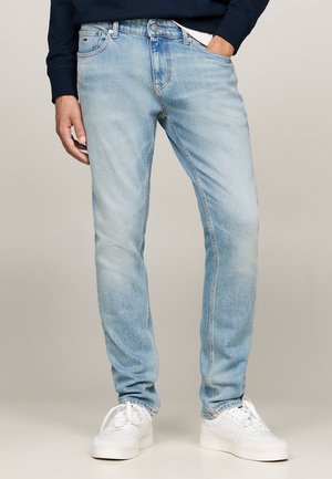 Lichtblauwe spijkerbroek gedragen met witte sneakers, één hand in de zak, gecombineerd met een donkerblauw shirt met lange mouwen.