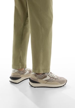 Sneaker beige con texture diverse, dettagli grigi, design con lacci e suola in gomma nera, abbinate a pantaloni verdi chiaro a sigaretta.
