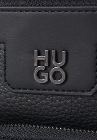 Pelle nera testurizzata con un logo "HUGO" in metallo argentato. Presenta un comparto con cerniera e bordi lisci.