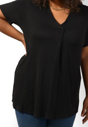 Nainen, jolla on päällään musta lyhythihainen V-pääntie paita, kultaketju kaulakoru ja siniset farkut, poseeraa yksi käsi lantiolla.