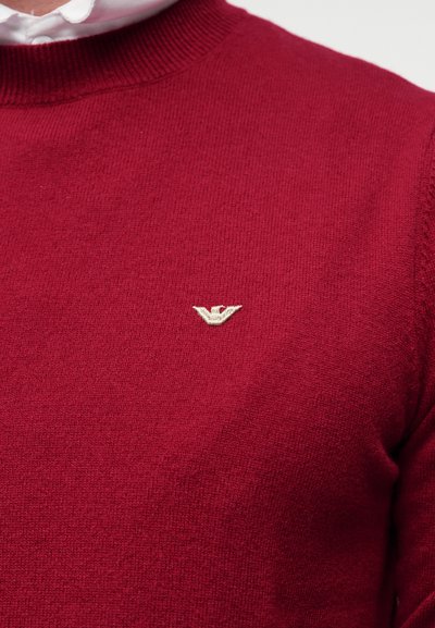 Κόκκινο πλεκτό πουλόβερ με στρογγυλή λαιμόκοψη, με μικρό μπεζ κεντητό λογότυπο στο στήθος, κατασκευασμένο από μαλακό, υφασμάτινο ύφασμα με υφή.