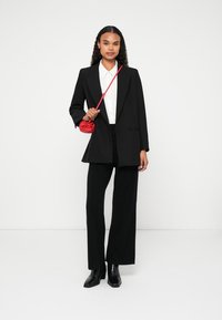 Černý oversized blazer přes bílou košili, doplněný o široké černé kalhoty a černé kotníkové boty. Drží malou červenou kabelku.