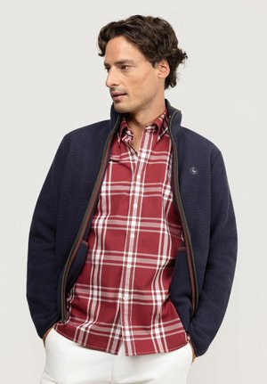 Hombre con chaqueta de forro polar azul marino sobre una camisa a cuadros roja y blanca con botones, con las manos en los bolsillos, mirando hacia un lado.