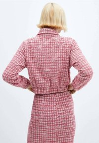 Personne aux cheveux blonds courts portant un manteau à carreaux roses et blancs ainsi qu'une jupe assortie, vue de dos, les mains sur les hanches.