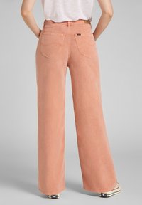 Jeans de pierna ancha en un suave color durazno, hechos de mezclilla con una textura suave. Presentan bolsillos traseros y detalles de costura sutiles.