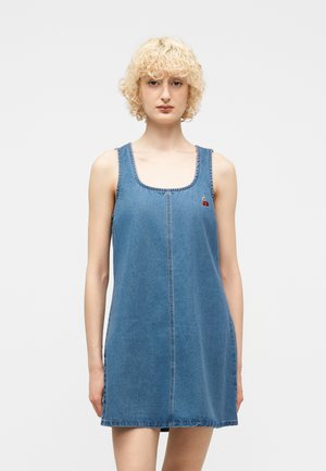 Blondynka w niebieskiej sukience jeansowej bez rękawów z małym haftem czereśni na lewej piersi, stojąca na jednolitym tle.