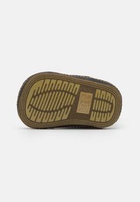Suela de un zapato marrón con patrones de tracción amarillos y un logo rectangular en el centro, sobre un fondo blanco liso.