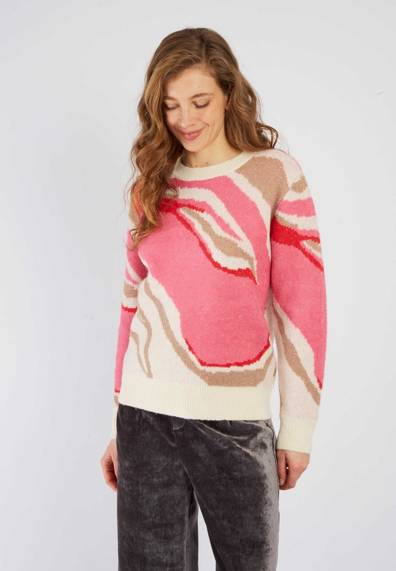 Pull en tricot avec un motif abstrait rose et crème, featuring une texture douce et un ourlet côtelé. Assorti avec un pantalon en velours gris.