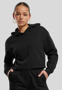 Juoda trumpa sudedamoji hoodie su raišteliu, ilgomis rankovėmis ir raukšlėtomis rankogaliais. Pagaminta iš minkštos medžiagos, pasižymi laisvu siluetu ir šoninėmis kišenėmis.