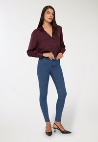 Kobieta poprawiająca guzik na bordowej jedwabnej bluzce, nosząca niebieskie wąskie dżinsy i czarne buty na obcasie typu slingback, stojąca na tle białego tła.