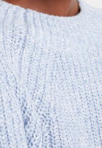 Hellblauer gestrickter Pullover mit einem strukturierten Muster und einem gerippten Kragen. Der Stoff wirkt weich und etwas voluminös.
