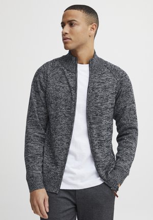 Chaqueta de punto gris con cremallera, patrón texturizado, puños acanalados y cuello alzado, llevada sobre una camiseta blanca.