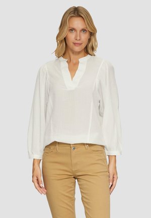 Weißes Blusenshirt mit strukturiertem Muster, V-Ausschnitt und langen Puffärmeln, kombiniert mit hellbraunen, figurbetonten Hosen.