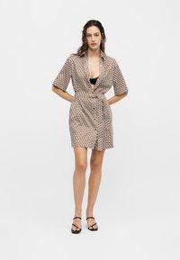 Γυναίκα στέκεται φορώντας ένα κοντό φόρεμα με μανίκια, με σχέδιο και ζώνη, μαύρο σουτιέν από κάτω και μαύρα ανοιχτά σανδάλια σε απλό λευκό φόντο.