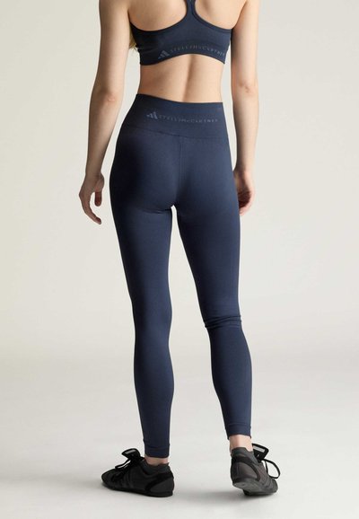 Frau trägt dunkelblaue, hoch taillierte Leggings und passenden Sport-BH mit schwarzen Sportschuhen, steht mit dem Rücken zur Kamera vor einfarbigem Hintergrund.