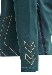 Teal sportjack met een ritszak, gouden geometrische patronen op de mouwen en zwarte accentvoering langs de zijkant.