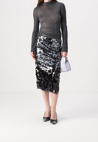 Jupe noire à sequins avec effet dégradé, associée à un haut gris transparent à manches longues et des talons noirs à bouts pointus. Petit sac à main blanc.