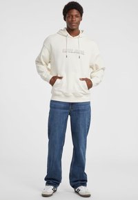 Sudadera crema con un bolsillo frontal y cordones, que presenta el texto "GUESS JEANS". Combinada con vaqueros azules y zapatillas blancas. Ajuste casual.