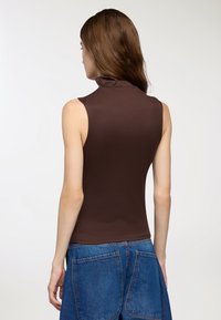 Brązowy bezrękawnik z wysokim kołnierzem i gładką fakturą, w połączeniu z dżinsową spódnicą z dużymi kieszeniami i luźnym krojem.