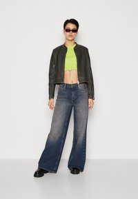 Veste en cuir noire courte, top court néon vert, jeans évasés bleus délavés et bottes noires chunky. Les accessoires comprennent des lunettes de soleil et une chaîne.