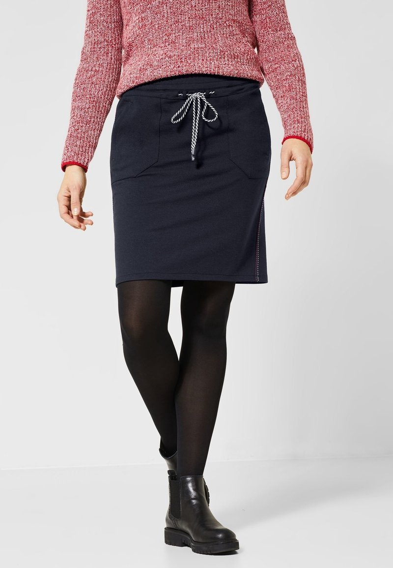 Tmavě modrá sukně s šňůrkou v pase, předními kapsami a bočním šitím. Kombinováno s černými punčochami a kotníkovými botami. Na vrchu texturovaný svetr.