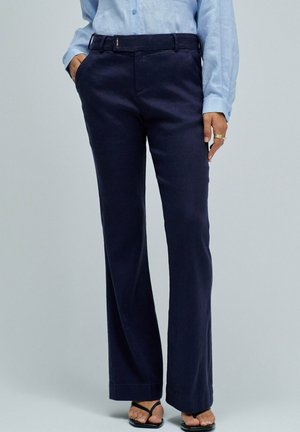 Personne portant un pantalon droit bleu marine, une chemise bleue claire boutonnée, des sandales noires à brides, avec la main dans la poche et des bagues aux doigts.
