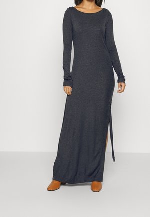 Femme portant une robe longue à manches longues gris foncé avec une fente latérale, associée à des chaussures marron à bout pointu, debout devant un fond uni.