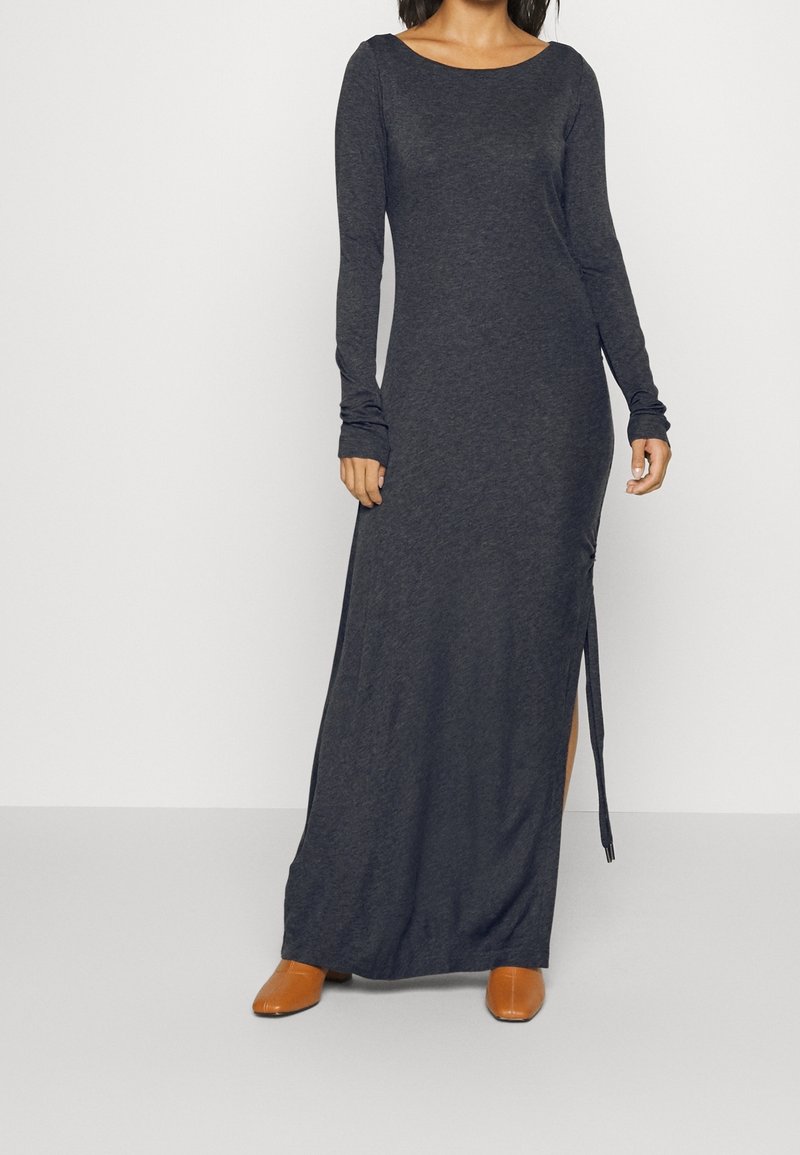 Femme portant une robe longue à manches longues gris foncé avec une fente latérale, associée à des chaussures marron à bout pointu, debout devant un fond uni.