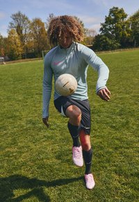 Athlet i lyseblå Nike langærmet trøje med rosa accenter, sorte shorts og rosa fodboldstøvler, der jonglerer med en fodbold på græs.