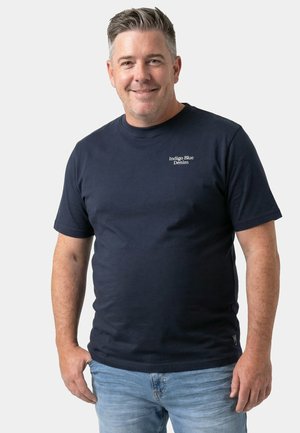 Lächelnder Mann mit kurzen grauen Haaren, der ein marineblaues „Indigo Blue Denim“-T-Shirt und hellblaue Jeans vor einem schlichten Hintergrund trägt.