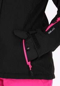 Gant noir avec fermeture éclair rose et bride velcro porté sur la main reposant sur la hanche, assorti à une veste noire et un pantalon rose vif.