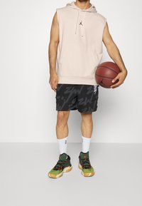 Beige sleeveless hoodie with front pocket, black shorts featuring a bold pattern, white socks, and green basketball sneakers with orange accents.