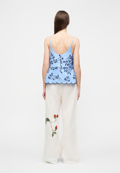 Γυναίκα με μακριά μαλλιά φορά μπλε αμάνικο τοπ με σκούρες ανθισμένες κεντήσεις και λευκό παντελόνι με φαρδιά πόδια με κόκκινες κεντήσεις φράουλας.