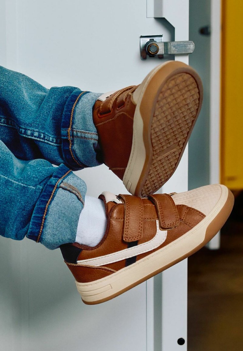 Enfant portant des baskets marron avec des chaussettes blanches et un jean bleu, pieds levés contre une porte blanche avec une serrure métallique.