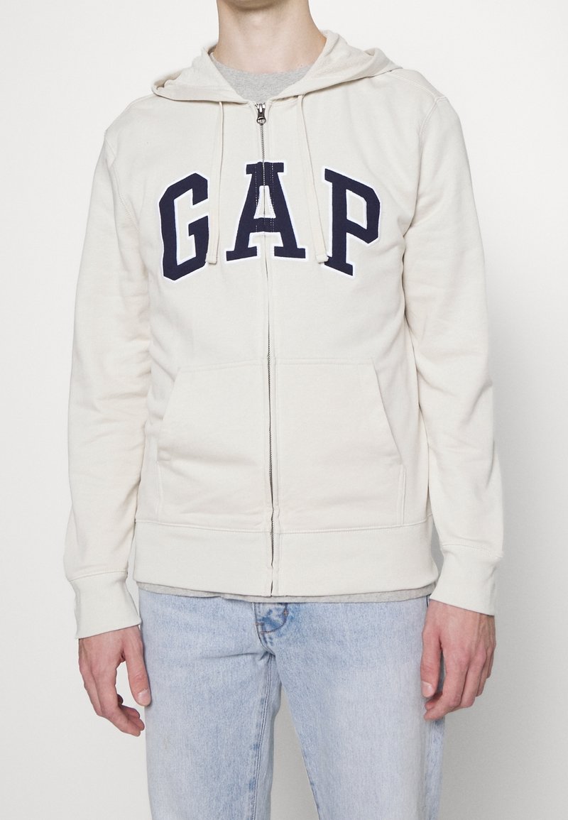 Felpa crema con zip e cappuccio, caratterizzata da un grande logo "GAP" in navy sul petto. Materiale morbido e due tasche laterali.