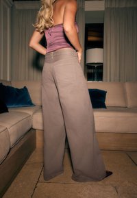 Jasnobrązowe spodnie z szerokimi nogawkami i dwoma tylnymi kieszeniami, wykonane z trwałej tkaniny. Modelka ma na sobie dopasowany fioletowy top i stoi obok kanapy.