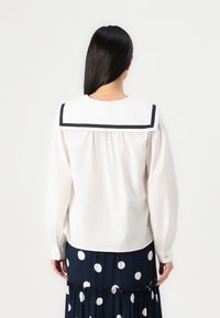 Hvit bluse med lange ermer og sjømannskrage, marinblå pynt og belagte mansjetter. Har en samlet bakdesign. Bæres med et marinblått prikkete skjørt.