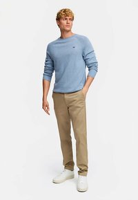 Lyseblå strikkegenser med rund hals, beige chinos og hvite joggesko. Genseren har en liten logo på brystet.