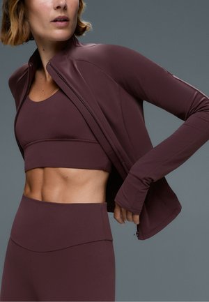 Donna che indossa un completo da allenamento viola scuro abbinato con top sportivo, leggings e giacca con zip su sfondo grigio uniforme.