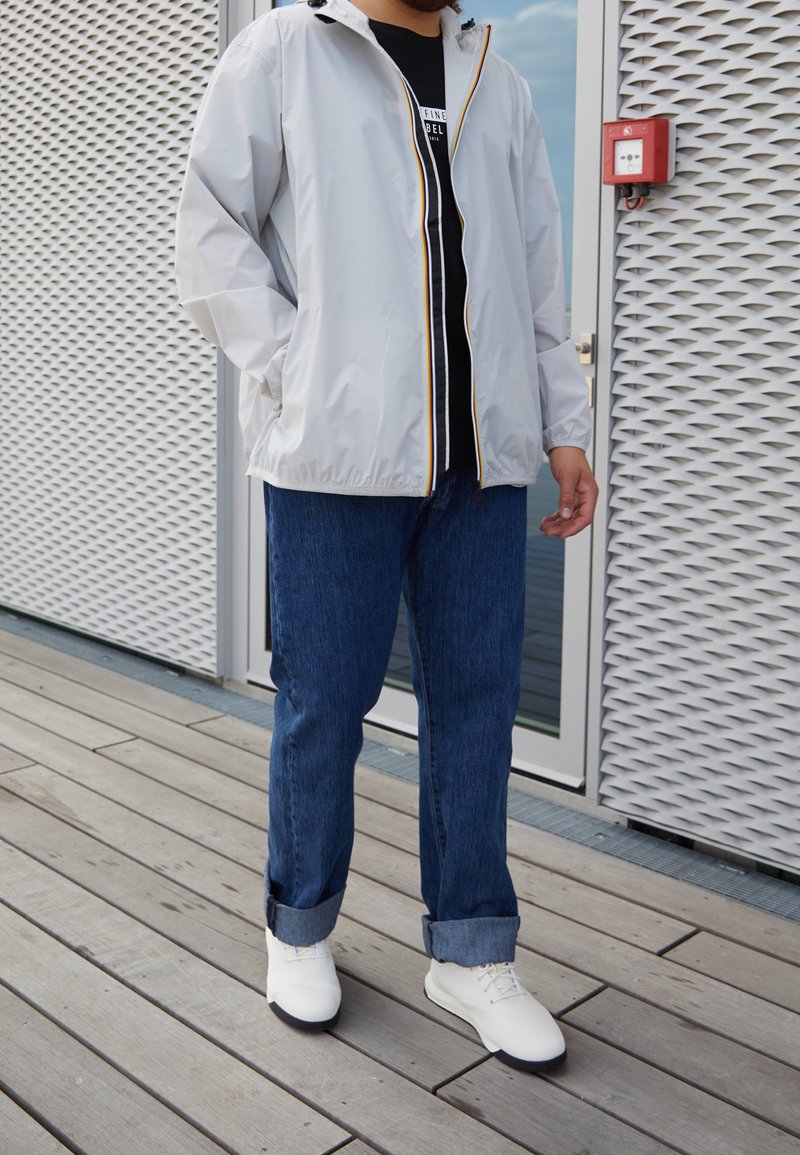 Ljusgrå zip-up jacka med gul kant, svart T-shirt, blå jeans och vita skor; avslappnad passform, slät textur och casual design.