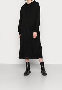 Haut à capuche noir court avec cordons de serrage, associé à une jupe midi plissée noire. Texture lisse et design ajusté. Porté avec des bottes noires à semelle épaisse.