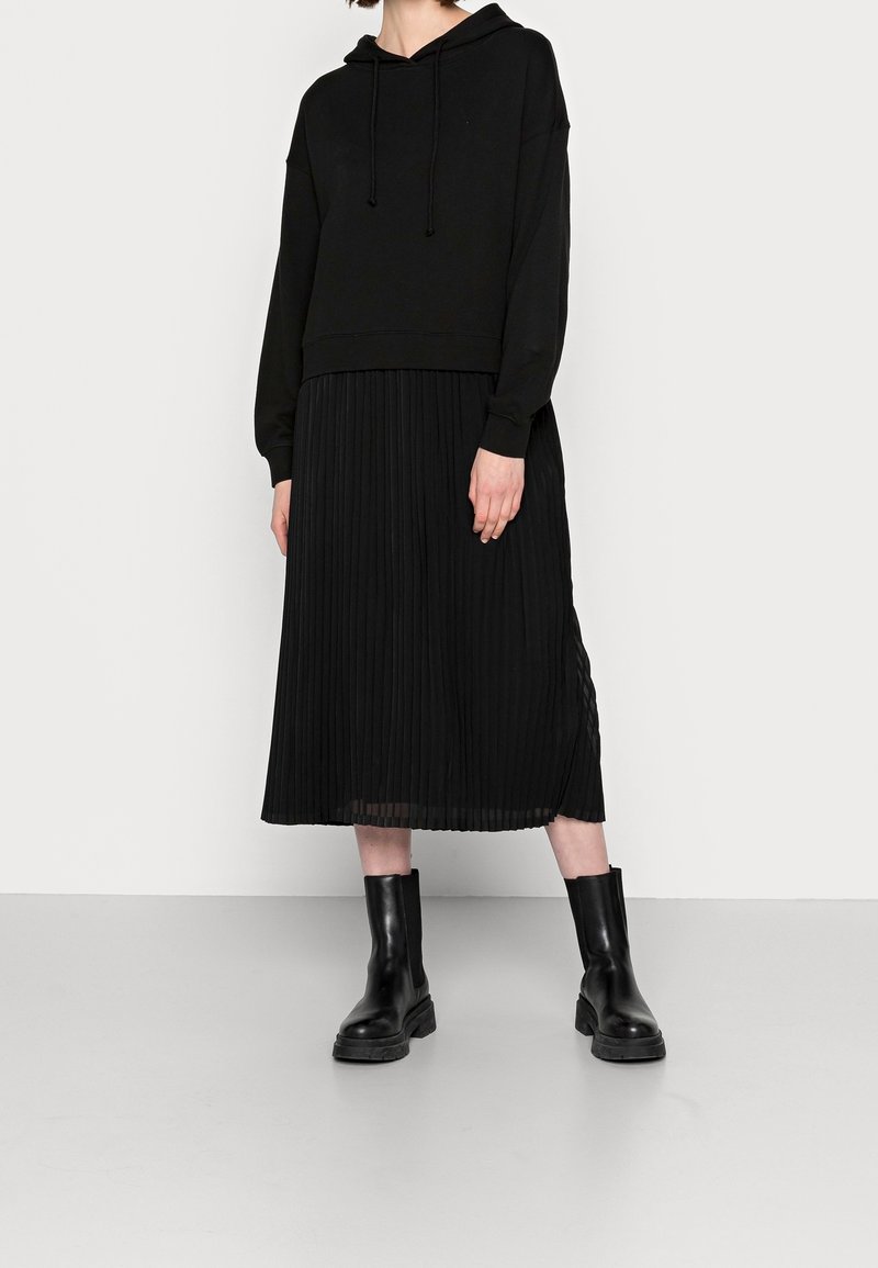 Haut à capuche noir court avec cordons de serrage, associé à une jupe midi plissée noire. Texture lisse et design ajusté. Porté avec des bottes noires à semelle épaisse.