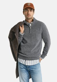 Grijze wollen trui met een rits tot de helft, gedragen over een gestreept shirt en gecombineerd met lichtblauwe jeans. Een bruine blazer hangt over de schouder.