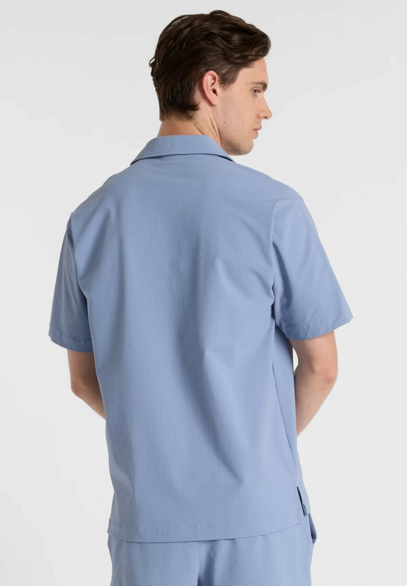 Chemise à manches courtes, bleu clair, en tissu texturé avec col cranté, présentant un ourlet droit et des fentes latérales pour le confort.
