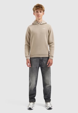 Tienerjongen staat rechtop met het gezicht naar voren, gekleed in een beige hoodie, vervaagde zwarte jeans en witte sneakers tegen een effen lichte achtergrond.