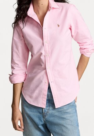 Femme portant une chemise rose clair à boutons avec les manches retroussées et un jean bleu, debout avec une main dans la poche.